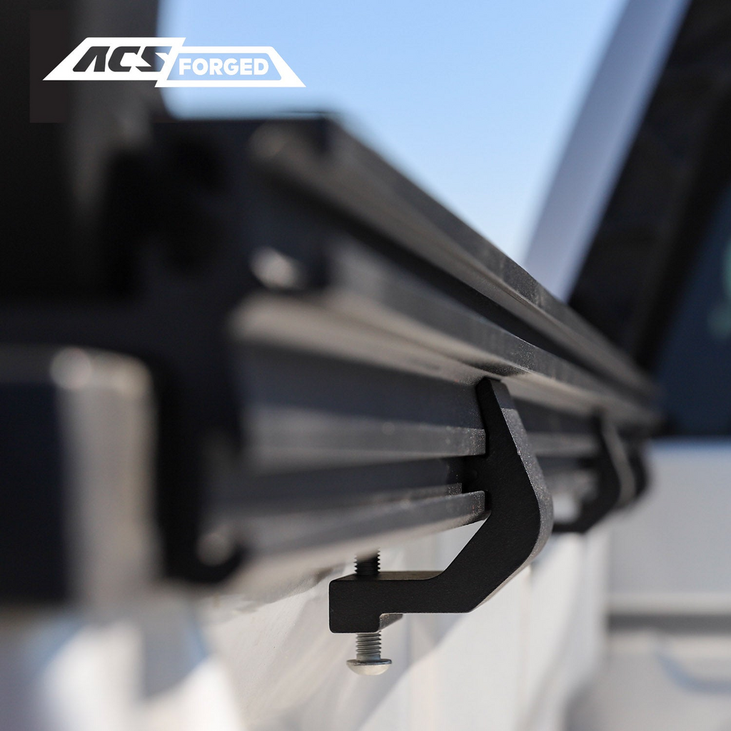 Close-up profile view of a black metal clamp secured to the underside of a truck bed rail, holding the ACS Forged base track in place.